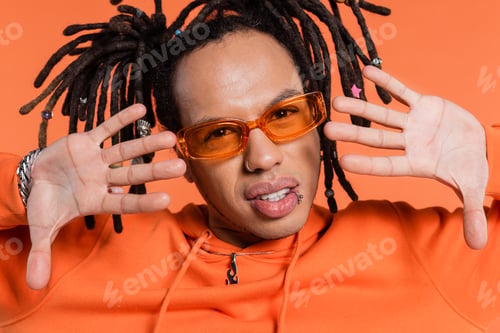 Preview: pierced multiracial man in hoodie and stylish sunglasses gesturing isolated on coral