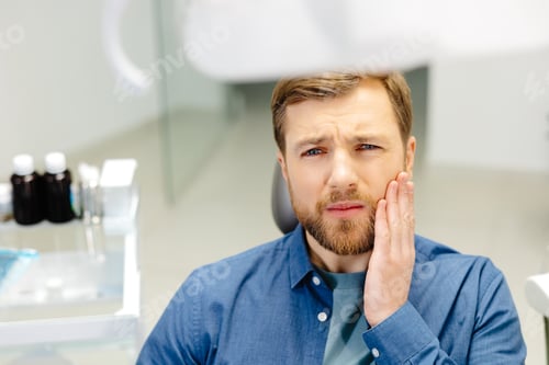 Preview: Sad man sitting in dental chair suffering from acute tooth pain, rubbing his cheek, close up