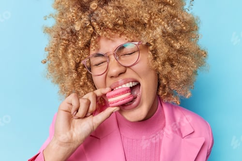 Preview: Portrait of lovely curly haired woman eats macaroon biscuit has sweet tooth enjoys unhealthy food we