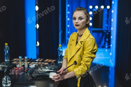 Preview: attractive ballerina in yellow leather jacket applying makeup in dressing room