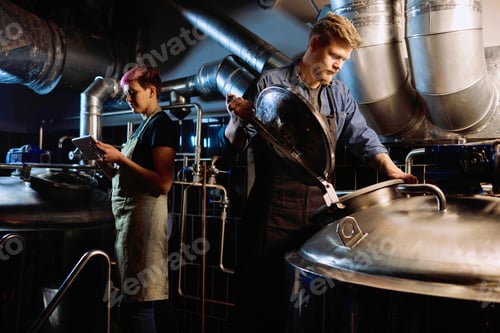 Preview: Young blond bearded male brewer in workwear opening lid of huge steel tank