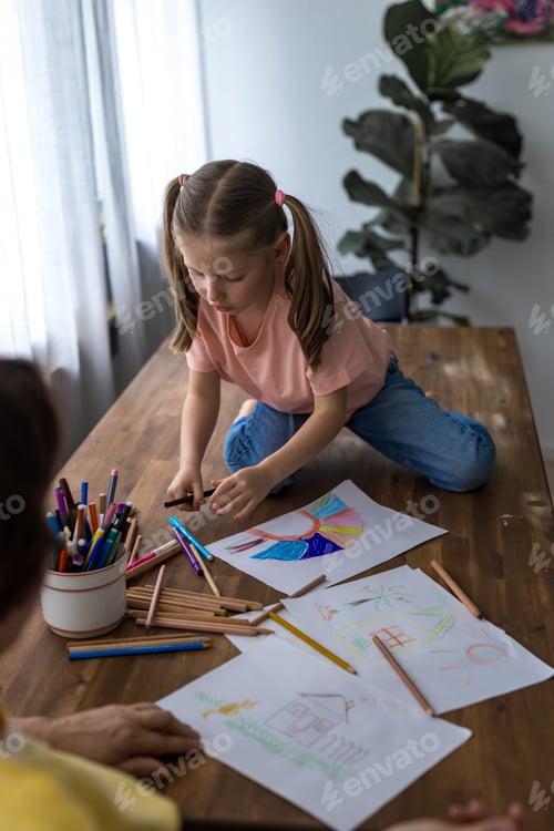 Preview: A 5-year-old girl laughs joyfully as she draws at the table, surrounded by colorful pencils