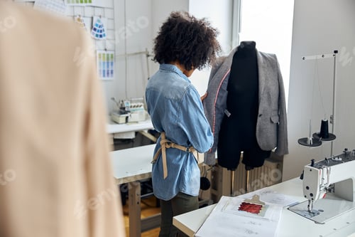 Preview: African-American lady tailor looks at new jacket on black mannequin in sewing workshop