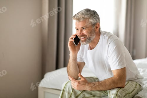 Preview: Happy middle aged man sitting on bed, have phone conversation