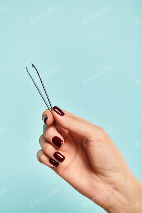 Preview: object photo of metal tweezers in hand of young unknown lady with nail polish on blue vivid backdrop