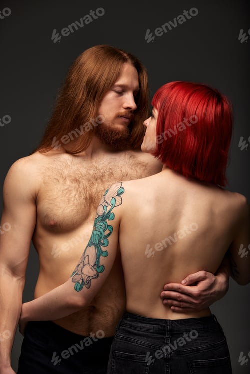 Preview: A young attractive woman with bright ruby hair, tattoos and piercings hugging a man with long hair