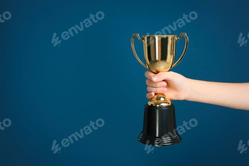 Preview: Child holding golden winning cup on dark blue background, closeup. Space for text