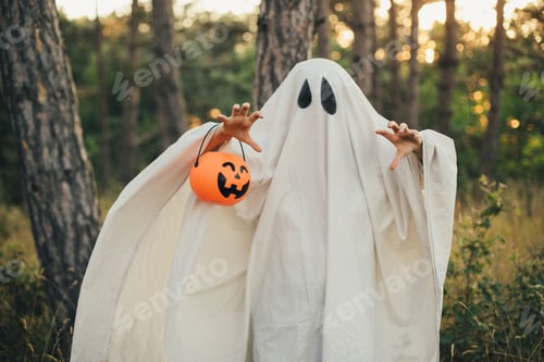 Preview: Girl wearing ghost costume holding pumpkin bucket with candies, standing in a forest.