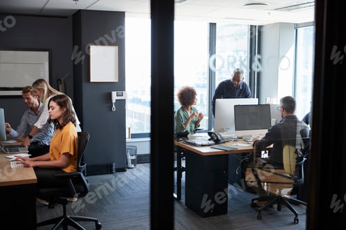 Visualização: Jovem equipe criativa trabalhando em conjunto em computadores em um escritório casual, vista através de uma parede de vidro