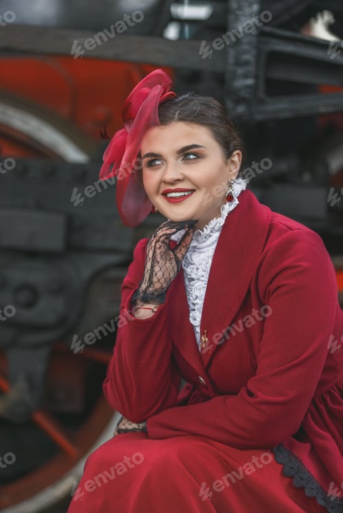 Preview: A girl in a burgundy suit is sitting on a suitcase near an old steam locomotive.Vintage portrait