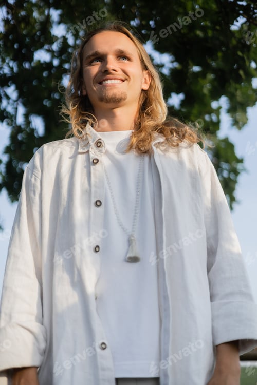Preview: low angle view of carefree long haired yoga man in linen clothes smiling outdoors
