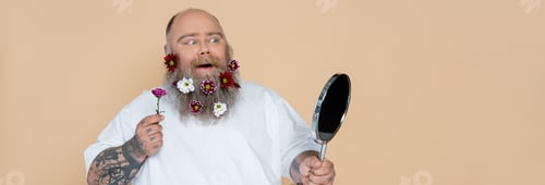 Preview: impressed overweight man with floral decor in beard looking in mirror isolated on beige, banner