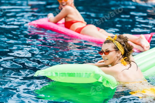 Preview: two attractive smiling women in swimsuits resting on inflatable mattresses in pool