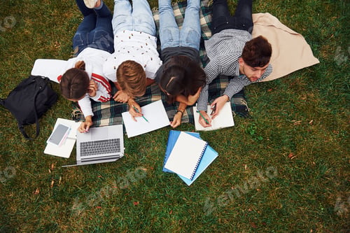 Preview: Writing on paper. With laptop. Group of young students in casual clothes on green grass at daytime