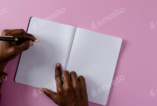 Preview: Hands of biracial man holding pen and writing in notebook with copy space on pink background