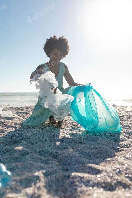 Preview: African american woman picking up garbage and collecting in plastic bag at beach against clear sky