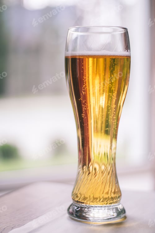 Preview: Glass of light beer with bubbles, on wooden table with white tablecloth near window. Vertical