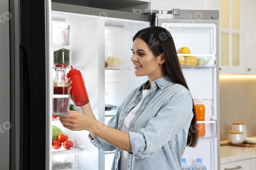 Preview: Young woman taking bottle of ketchup out of refrigerator in kitchen