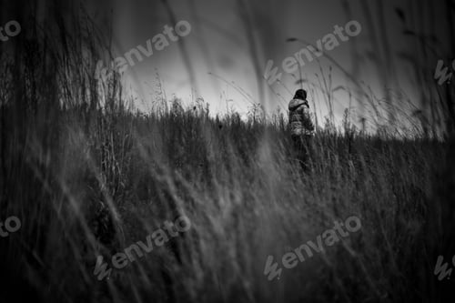 Preview: Grayscale shot of a person walking in a field covered in the grass under a cloudy sky