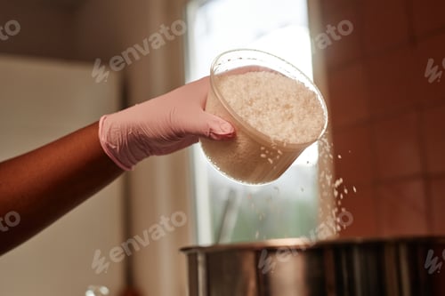 Preview: Hand of Black Woman in Glove Pouring Coconut Flakes Making Bread Dough in Bakery Kitchen