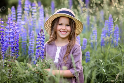 Preview: little caucasian girl in a linen dress and a straw hat in a field with lupins flowers