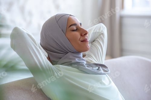 Preview: Young muslim woman resting at home with eyes closed hands behind head napping sitting on sofa