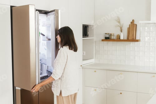 Preview: Stylish woman opening fridge and looking inside in new minimal white kitchen
