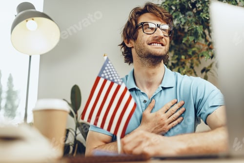 Preview: Young bearded businessman sitting at table with laptop and american flag