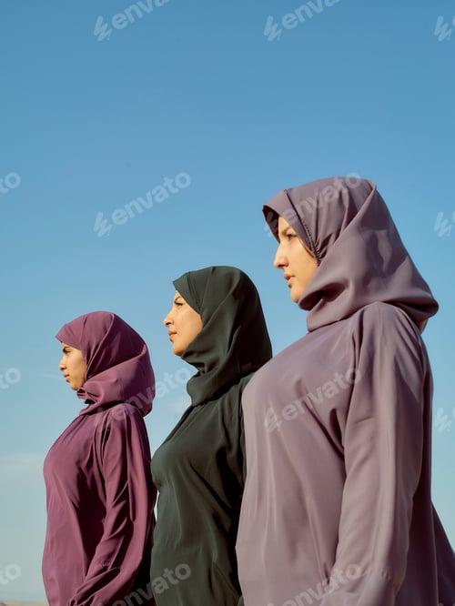 Preview: Three women wearing hijabs standing in profile against a clear blue sky.