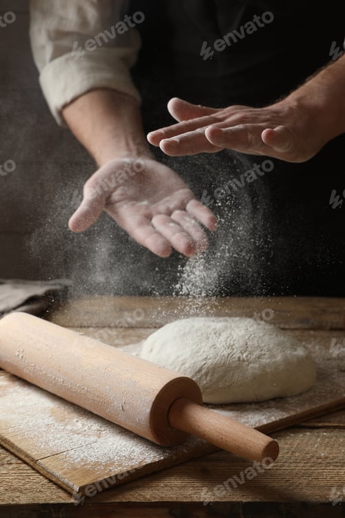 Preview: Man sprinkling flour over dough at wooden table, closeup