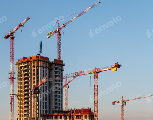 Preview: several construction cranes stand next to a tall skyscraper and one of them has yellow lights