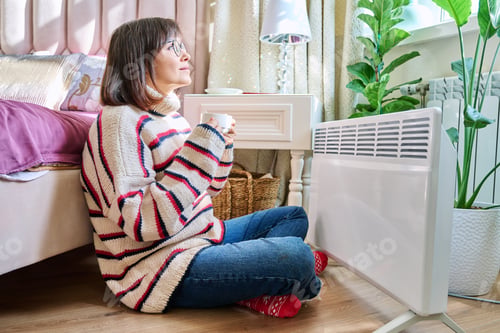 Preview: Woman warming herself in bedroom near electric heater
