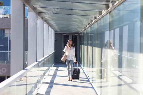 Preview: Young biracial businesswoman talking on phone while walking with luggage at airport corridor