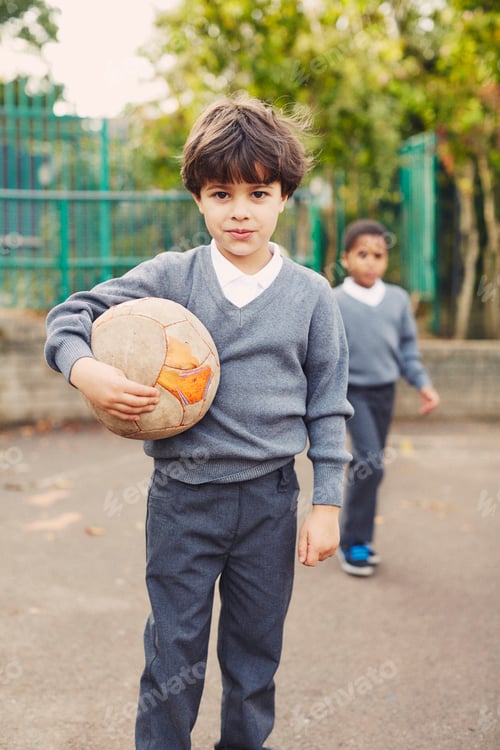 Preview: Young boy confidently holding a soccer ball with a friend in the background at a schoolyard.