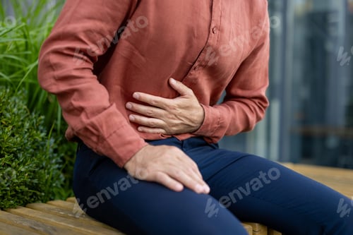 Preview: Man in casual attire sitting outdoors holding stomach showing symptoms of discomfort possibly