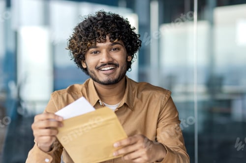 Preview: Young cheerful businessman with curly hair holding an envelope indoors