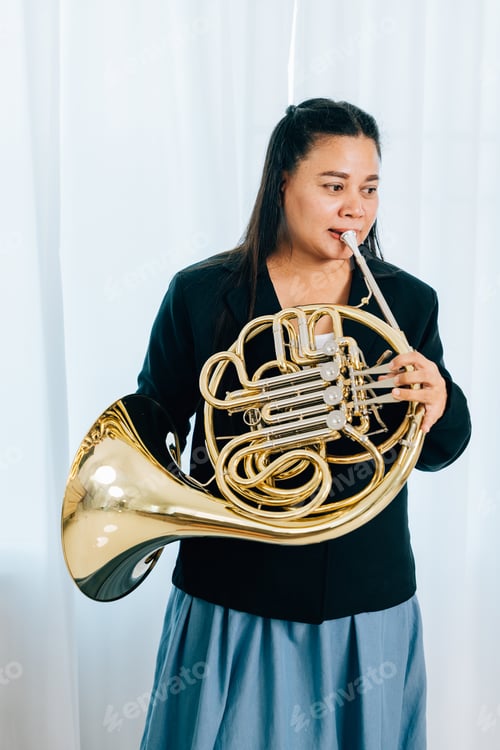 Preview: A woman stands at home in front of elegant curtains, playing a shiny gold horn. The warm lighting