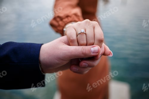 Preview: Image of a man putting a silver wedding ring on a woman's hand, outdoors. Sea or river background