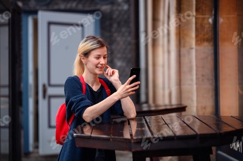 Preview: Young blonde woman in autumn-spring outwear and red backpack stands near high wooden table