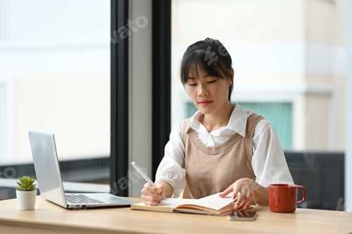 Preview: Focused asian female student taking notes while using laptop computer at coffee shop