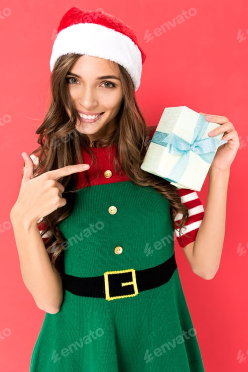 Preview: portrait of smiling woman in elf costume pointing at christmas gift isolated on red