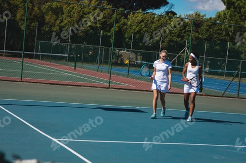 Preview: Smiling multiracial female players talking while walking at tennis court on sunny day