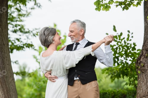 Preview: smiling mature man in formal wear dancing with bride in white wedding dress in green garden