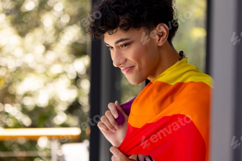 Preview: Smiling young transgender woman holding rainbow flag, celebrating LGBTQ+ pride at home