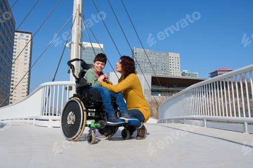 Preview: Mother holding hands with son with disability in wheelchair on bridge - cerebral palsy concept
