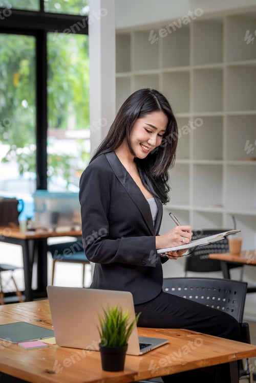 Preview: Side view of young Asian businesswoman holding pen a taking note sitting at the edge of a laptop tab