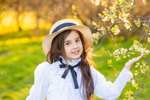 Preview: a adorable girl in a hat and white openwork dress, holding a branch of a tree blooming with white fl