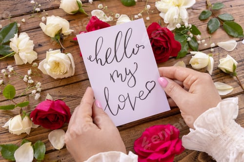 Preview: Hands with card HELLO MY LOVE near red and cream flowers close up on a wooden table
