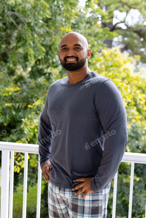 Preview: Portrait of happy biracial man in pyjamas standing on sunny balcony at home in front of treetops