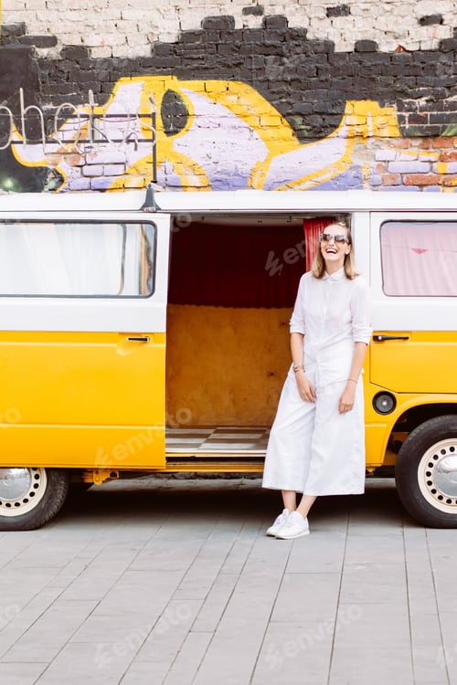 Preview: young fashionable woman in a white outfit near a yellow photo booth minibus on the street in summer